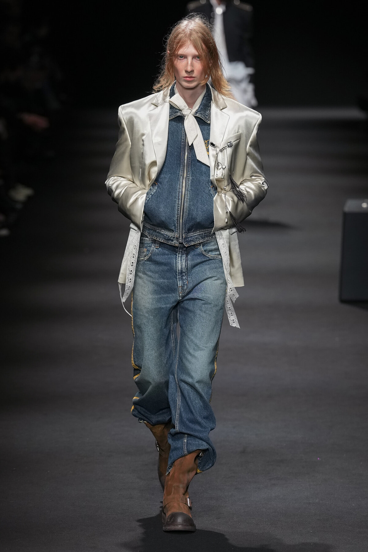 Female model on a fashion runway wearing a metallic silver oversized blazer over a denim jacket and jeans, with a white neck scarf and brown boots.