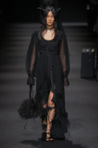 Model walks a dark runway in a long black dress with sheer sleeves and feathered accents, carrying a black feathered handbag.