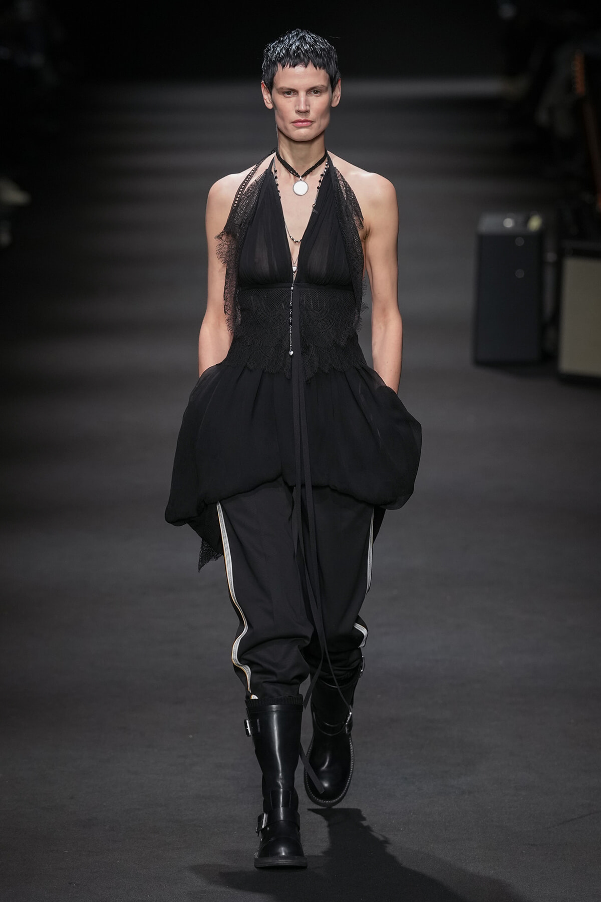 Model walking a runway in a black avant-garde dress with lace bodice and layered skirt, paired with tall black boots.