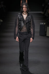 Model walks a dark runway in a sheer black lace top under a cropped black leather vest with silver buttons and wide-leg trousers.