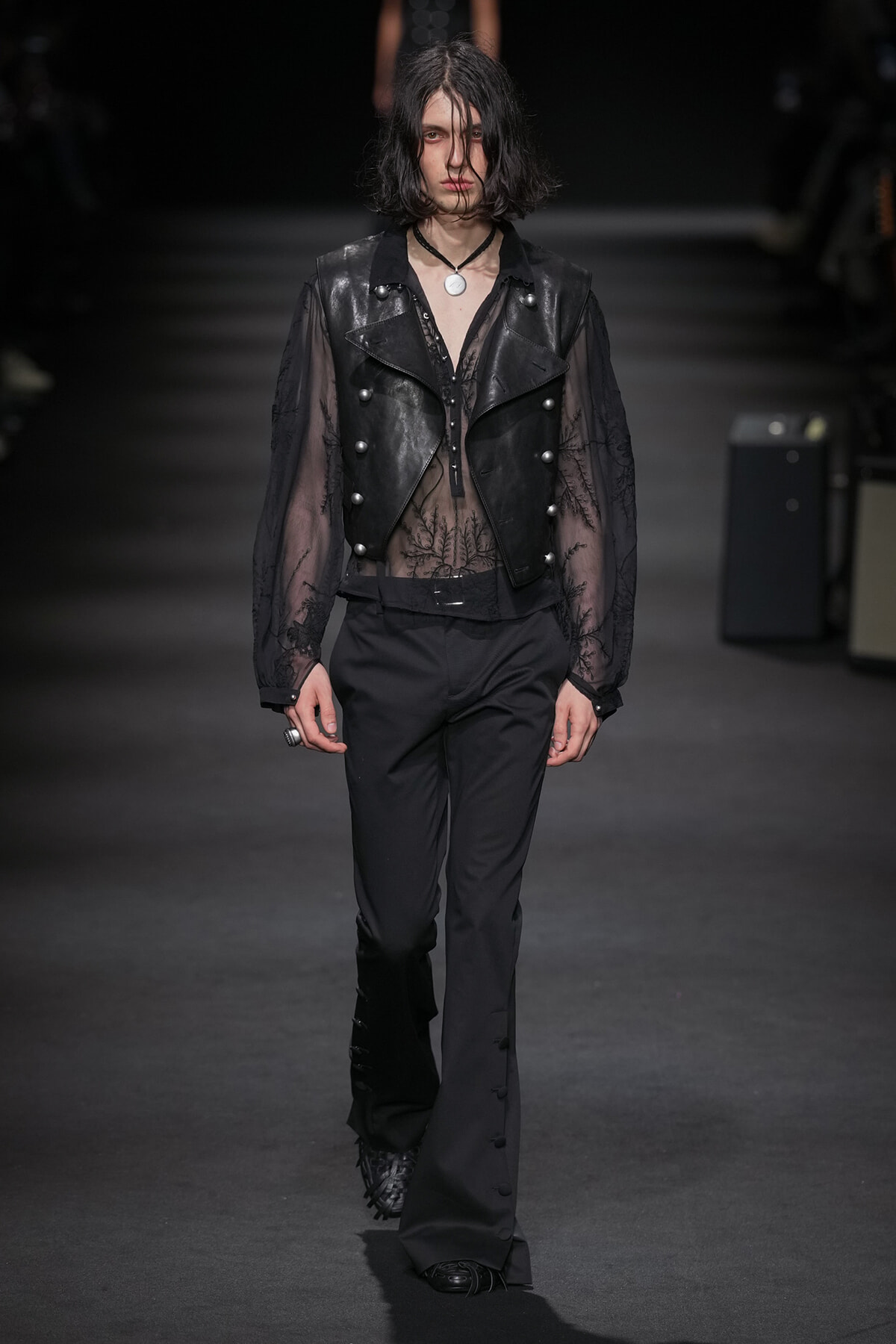 Model walks a dark runway in a sheer black lace top under a cropped black leather vest with silver buttons and wide-leg trousers.