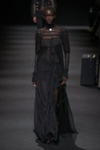 Model walks a dark fashion runway in an all-black lace and sheer gown with layered panels and tied belt, spotlight on her.