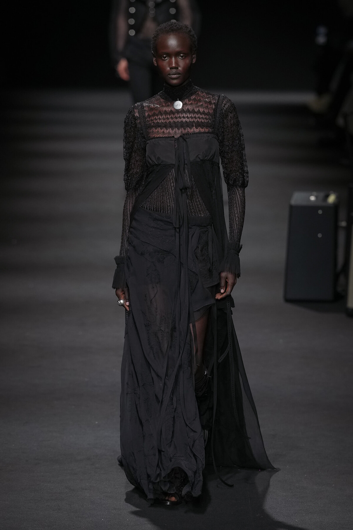 Model walks a dark fashion runway in an all-black lace and sheer gown with layered panels and tied belt, spotlight on her.