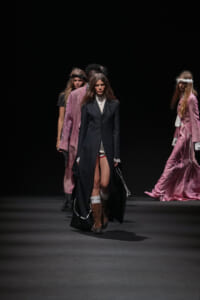 Models walk a fashion show on a dark runway; foreground model wears a long black coat, white blouse, and brown knee-high boots while other models in pink outfits stand in the background.