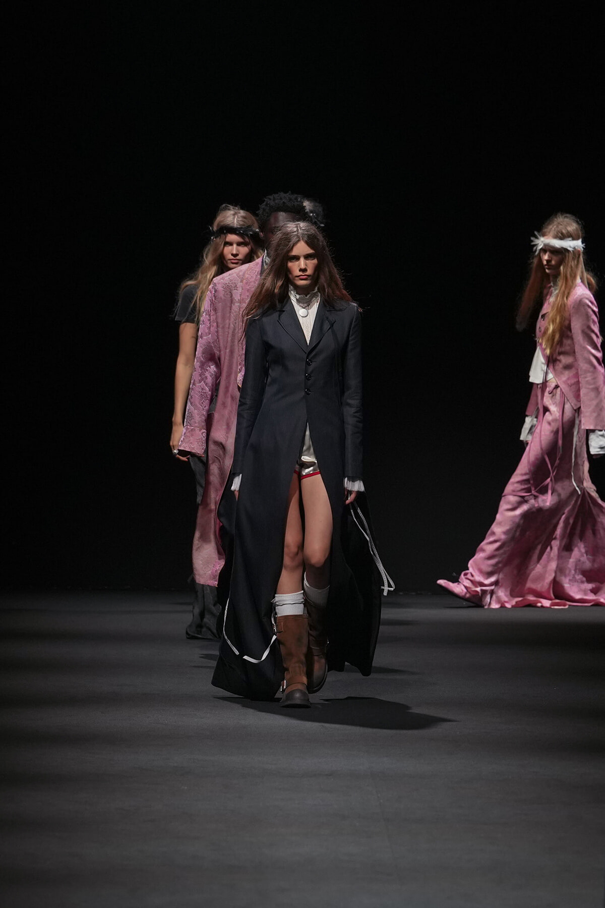 Models walk a fashion show on a dark runway; foreground model wears a long black coat, white blouse, and brown knee-high boots while other models in pink outfits stand in the background.