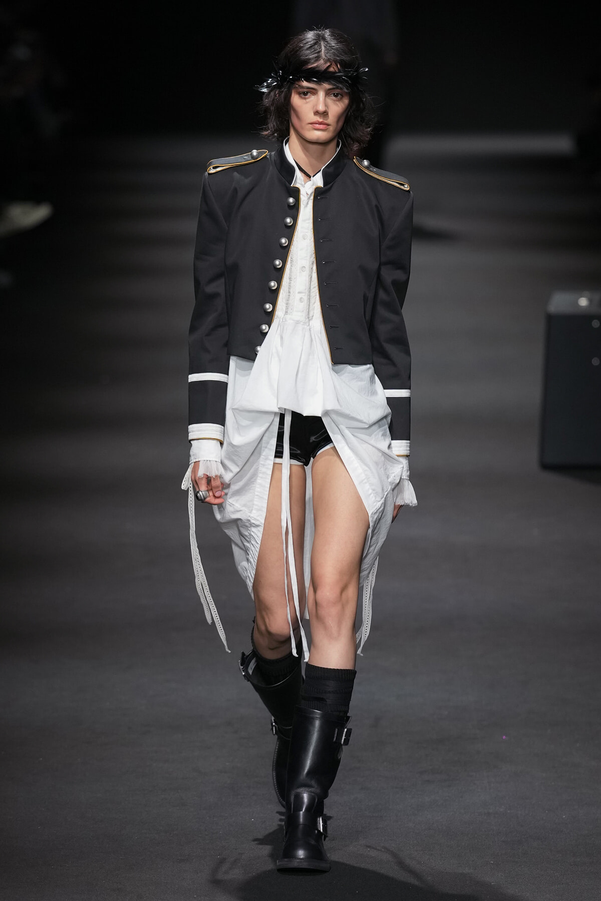Model on a runway wearing a black military-style jacket with gold epaulets, white shirt, and draped white skirt over black shorts, with black boots.