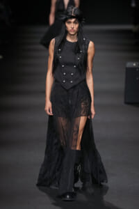 Model walking a dark runway in an all-black outfit: sleeveless vest with metal buttons over a sheer lace skirt and tall boots.