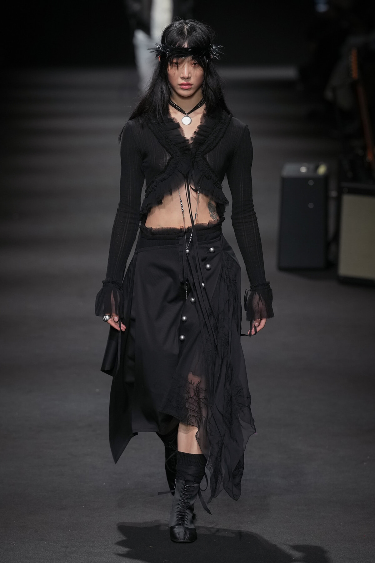 Model walking a fashion runway in a black Gothic-inspired outfit with a cropped top and long skirt, headband, and boots.