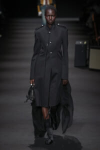 Full-length shot of a model walking the runway in a tailored black coat with a high collar, metal buttons, and epaulets, carrying a small black handbag and wearing lace-up boots.