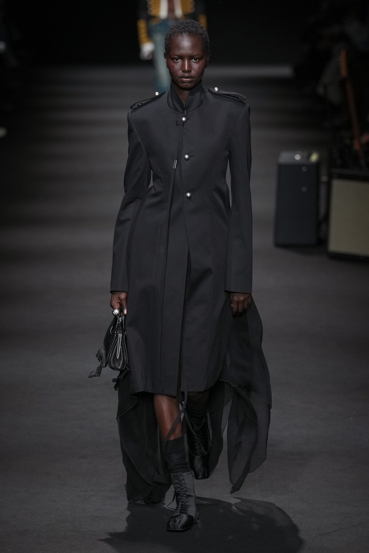 Full-length shot of a model walking the runway in a tailored black coat with a high collar, metal buttons, and epaulets, carrying a small black handbag and wearing lace-up boots.