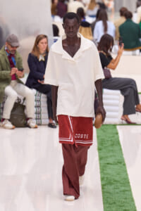 Male model walks a runway in a loose white polo and red textured wrap with 'COSTE' lettering, holding a small brown clutch.