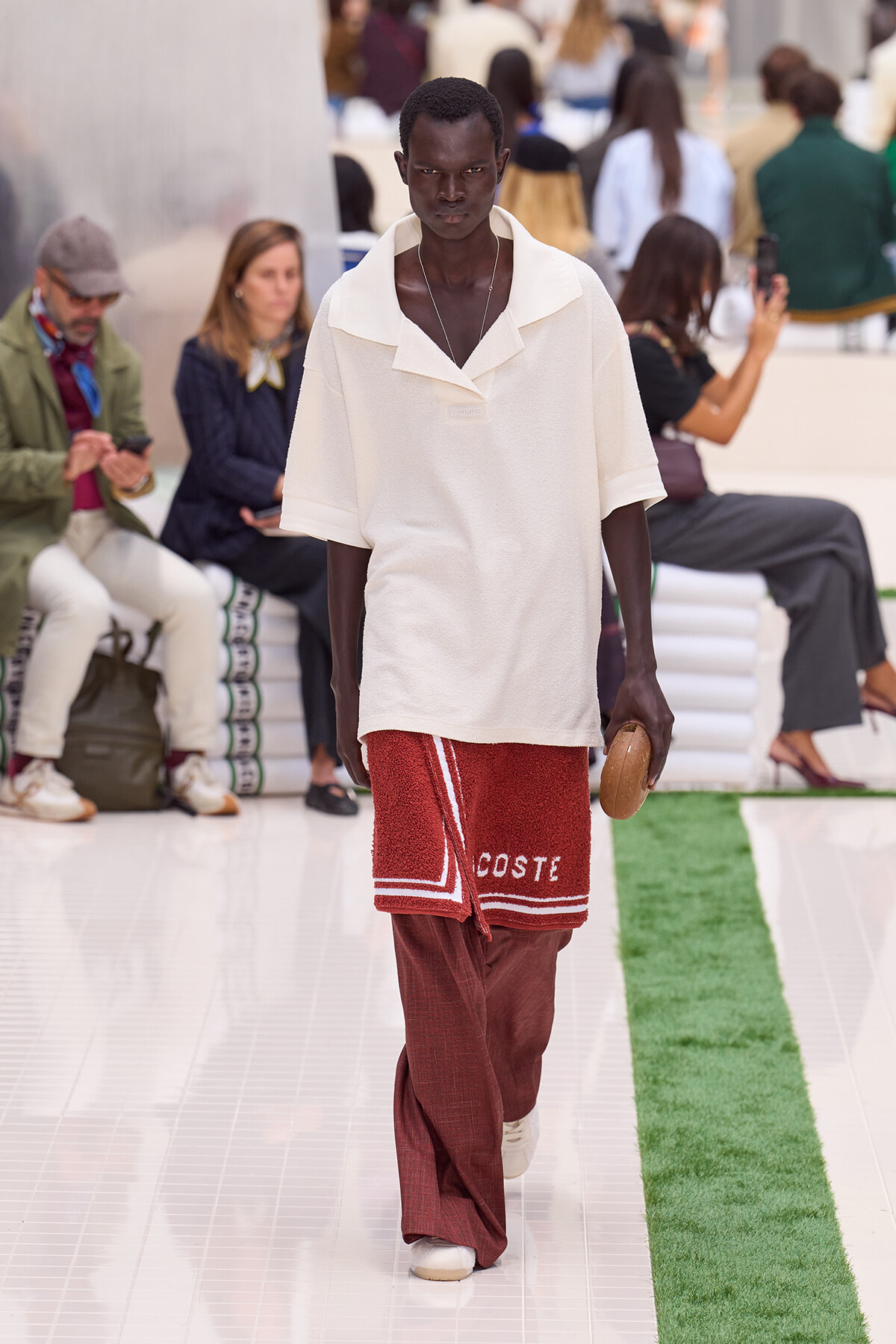 Male model walks a runway in a loose white polo and red textured wrap with 'COSTE' lettering, holding a small brown clutch.