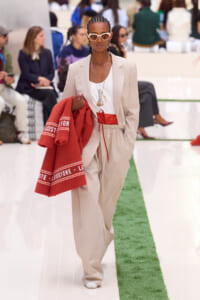 Model walks a fashion runway in a beige suit with a white top and red drawstring waist, accessorized with white sunglasses and layered necklaces, carrying a red towel.