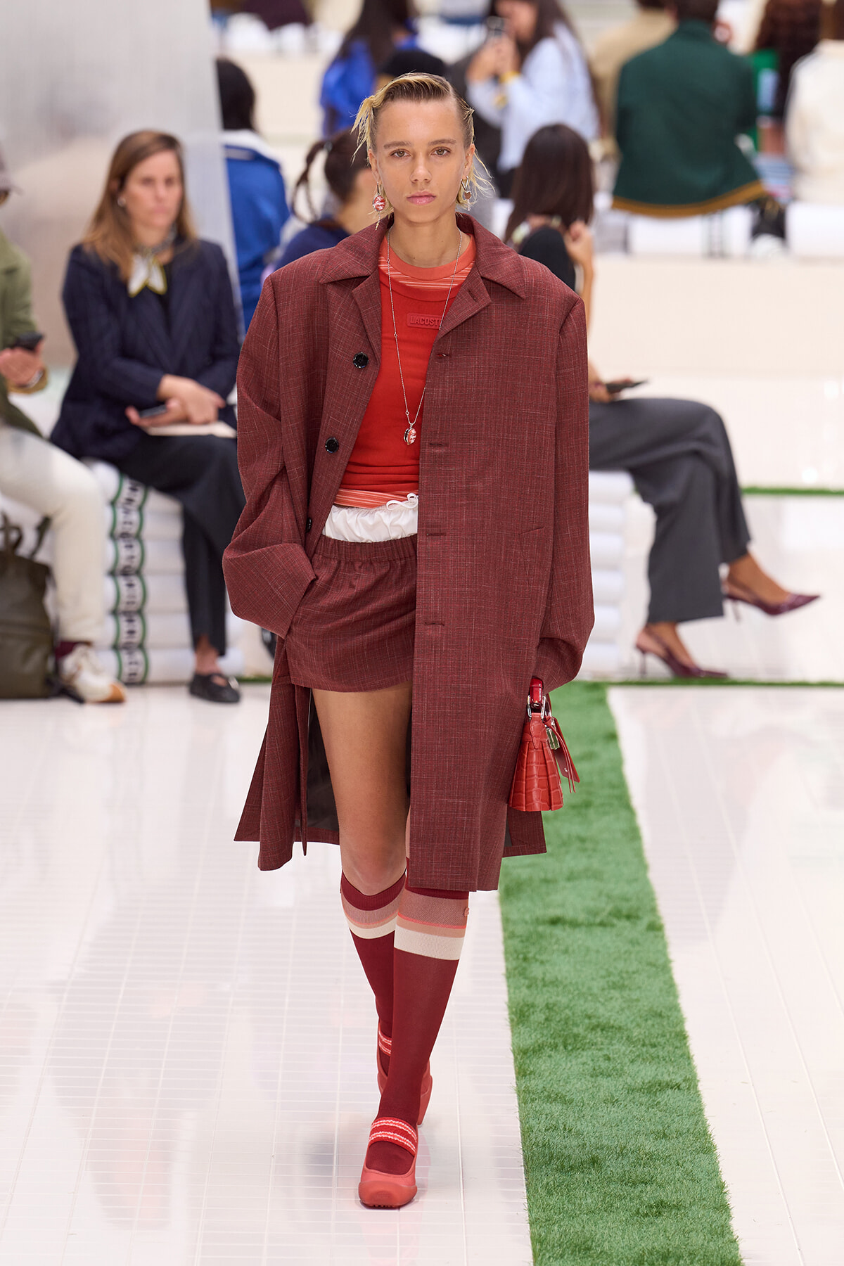 Model on a runway wearing a burgundy coat over a red outfit with matching socks and red bag, audience in the background.