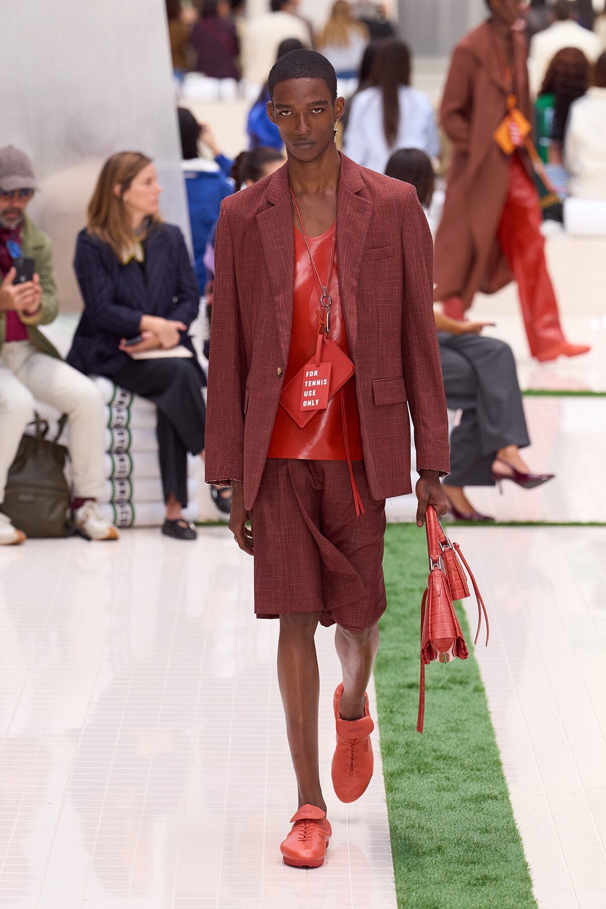 Male model walks the runway in a red-toned suit, matching red shirt, and red accessories, carrying a red handbag; tag on the outfit reads 'FOR TENNIS USE ONLY'.
