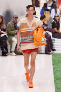Male runway model wearing a beige striped knit top, red shorts, and bright orange sneakers, carrying an orange backpack as he walks the catwalk.