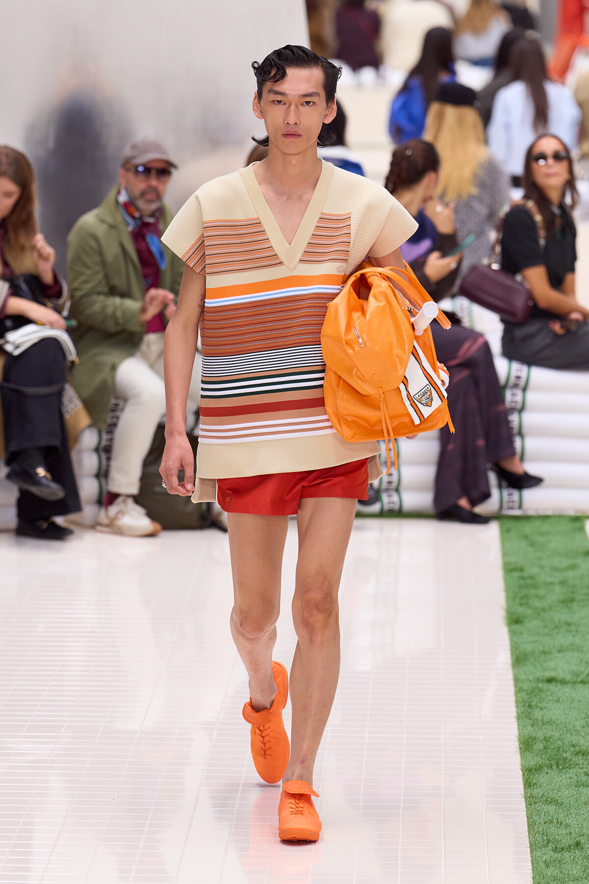 Male runway model wearing a beige striped knit top, red shorts, and bright orange sneakers, carrying an orange backpack as he walks the catwalk.