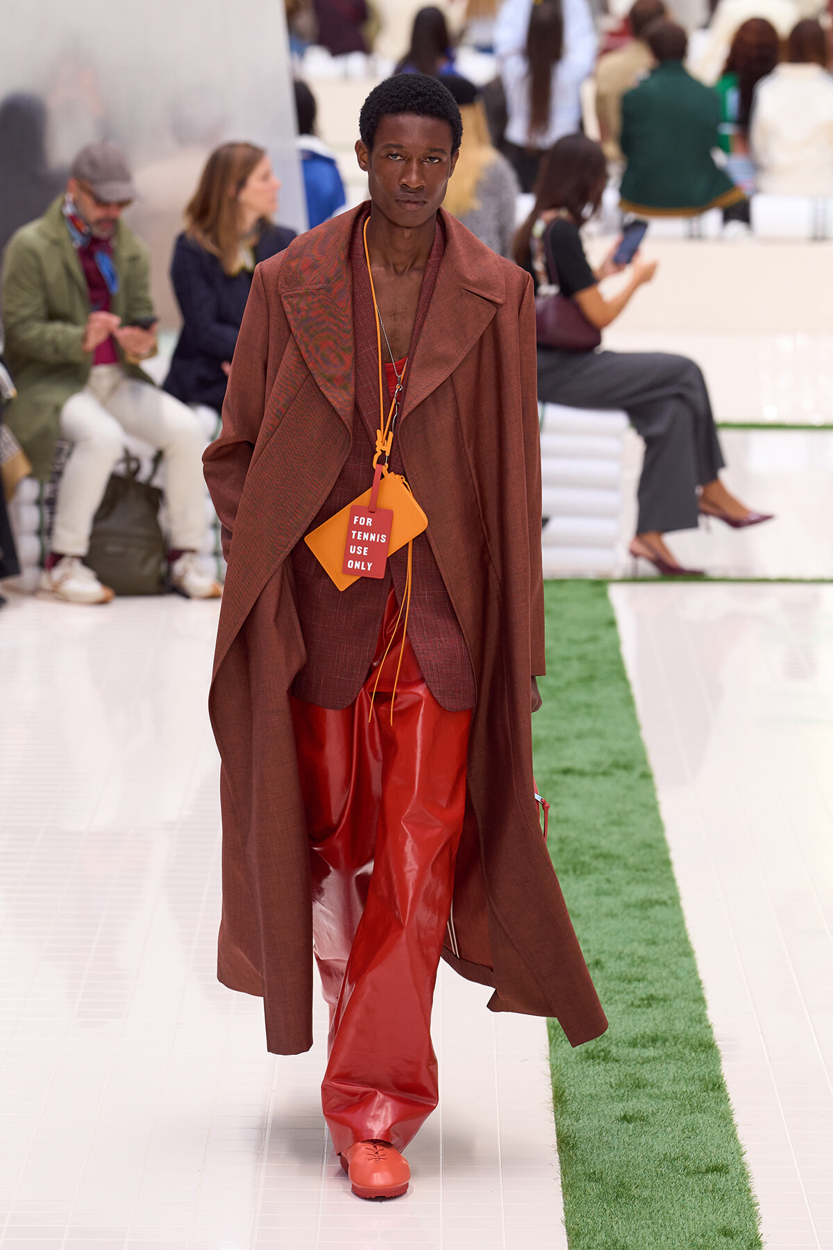 Male model on a fashion runway wearing a brown overcoat and red outfit with an orange tag that reads 'FOR TENNIS USE ONLY' hanging at chest level.