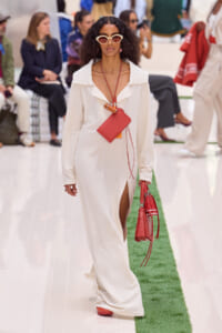 Model on a runway wearing a white textured maxi dress with a thigh-high slit, oversized collar, cream sunglasses, and red handbag and shoes with layered necklaces.