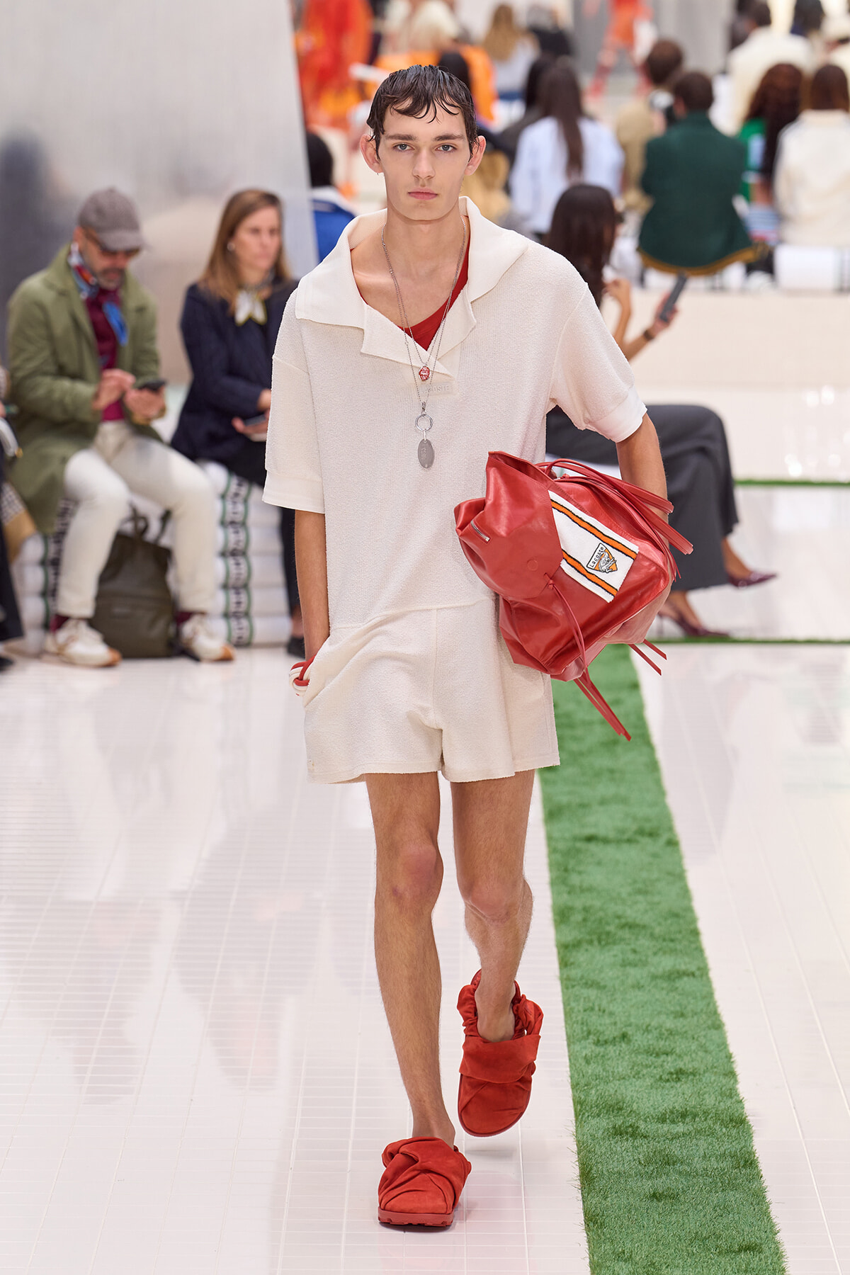 Male model walking a fashion runway in a cream textured polo and shorts, carrying a red bag under his arm and wearing red slide sandals.