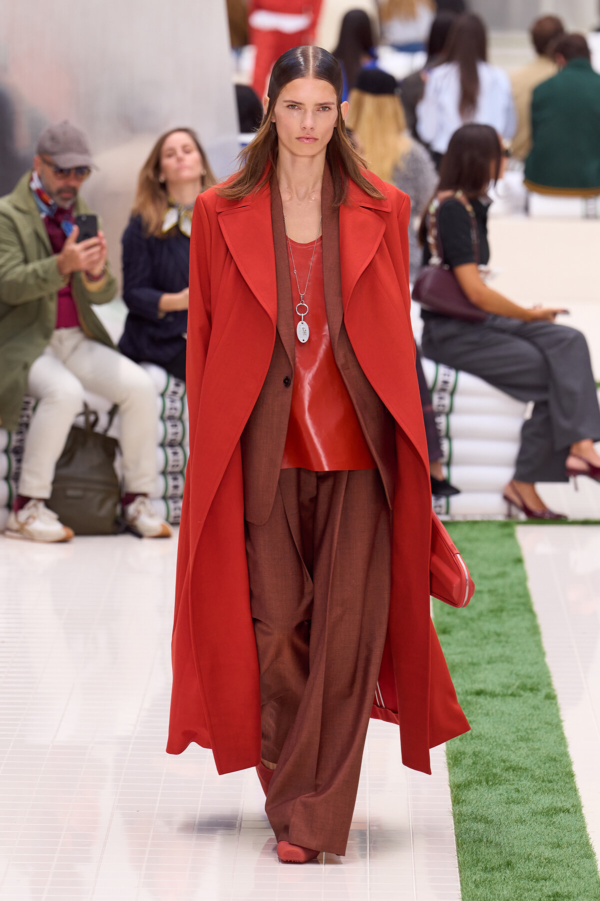 Model walks the runway in a bold red overcoat over brown suit and red top, accessorized with layered necklaces.