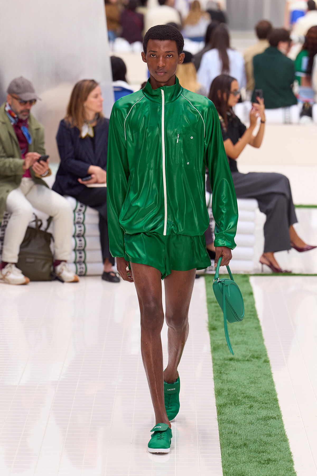 Male model walks the runway in a bright green tracksuit with white zipper, carrying a matching green bag on a glossy white catwalk.