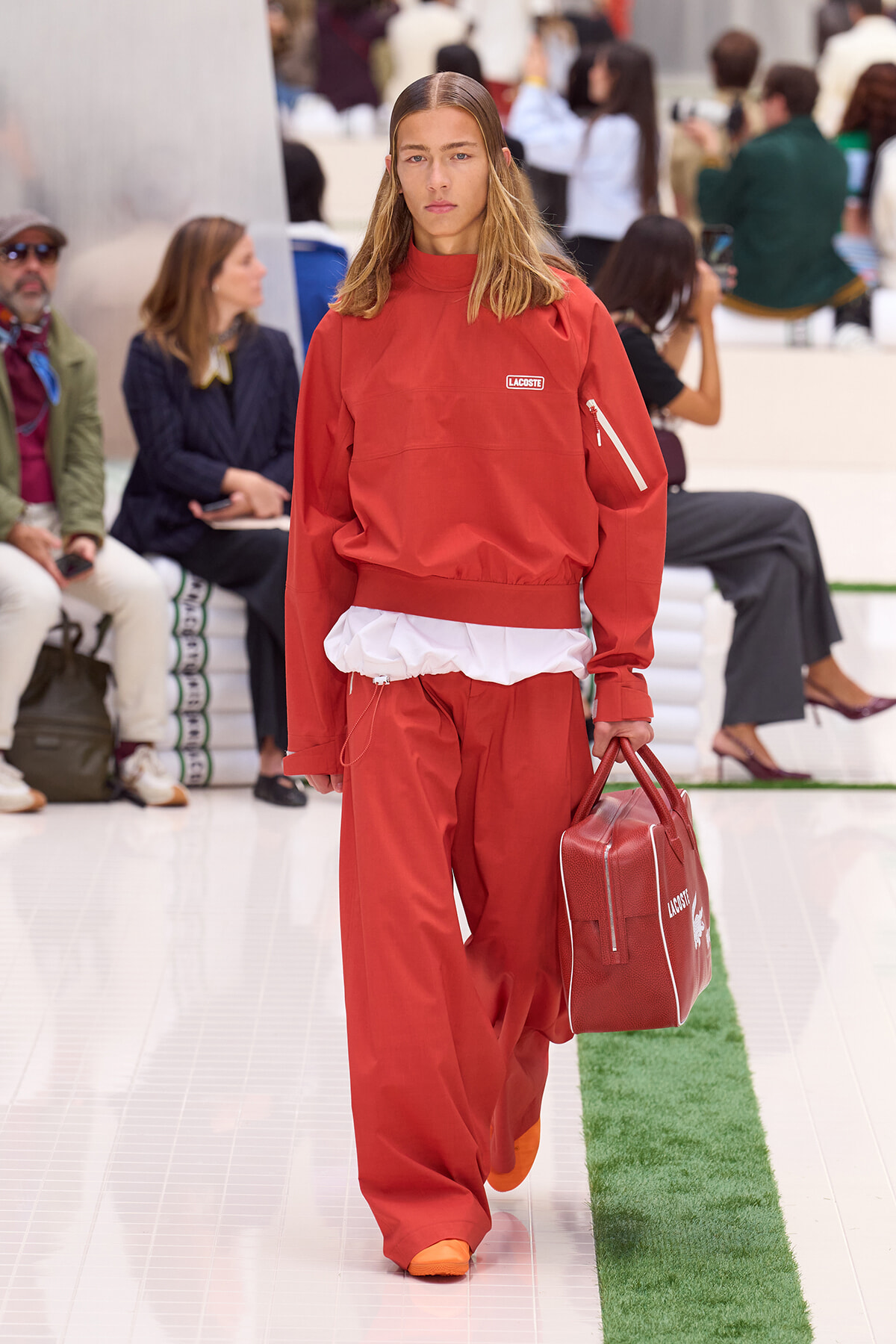 Model walks a fashion runway in a red Lacoste tracksuit, white peeking layer at the waist, carrying a red structured bag.