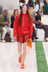 Model walking a fashion show runway in a red zip-up windbreaker, red shiny shorts, and bright orange sneakers, carrying an orange drawstring bag; audience sits in the background.