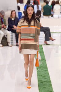 Model walking a fashion runway in a color-block striped top and beige skirt, carrying an orange handbag.