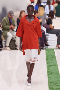 Model walks the runway in a loose red top paired with a white textured knee-length skirt with red trim and lettering, white shoes