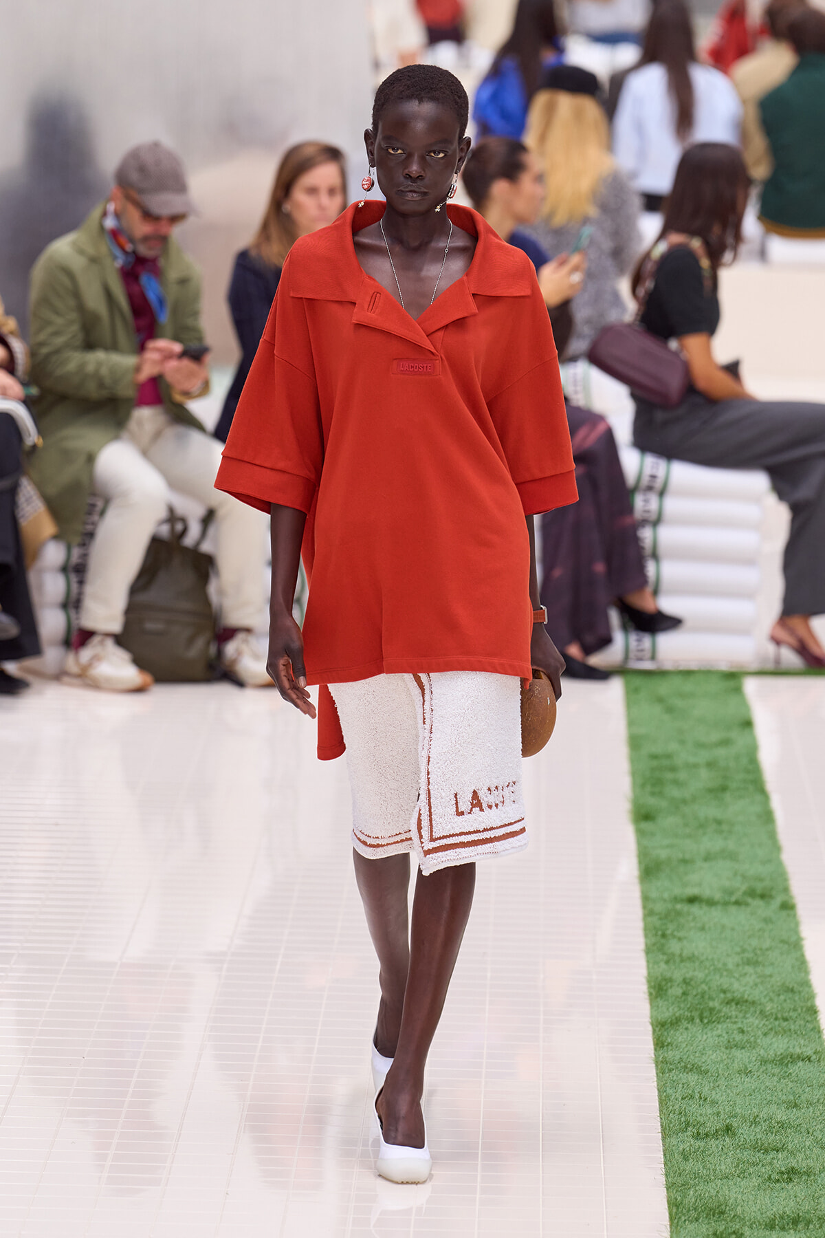 Model walks the runway in a loose red top paired with a white textured knee-length skirt with red trim and lettering, white shoes