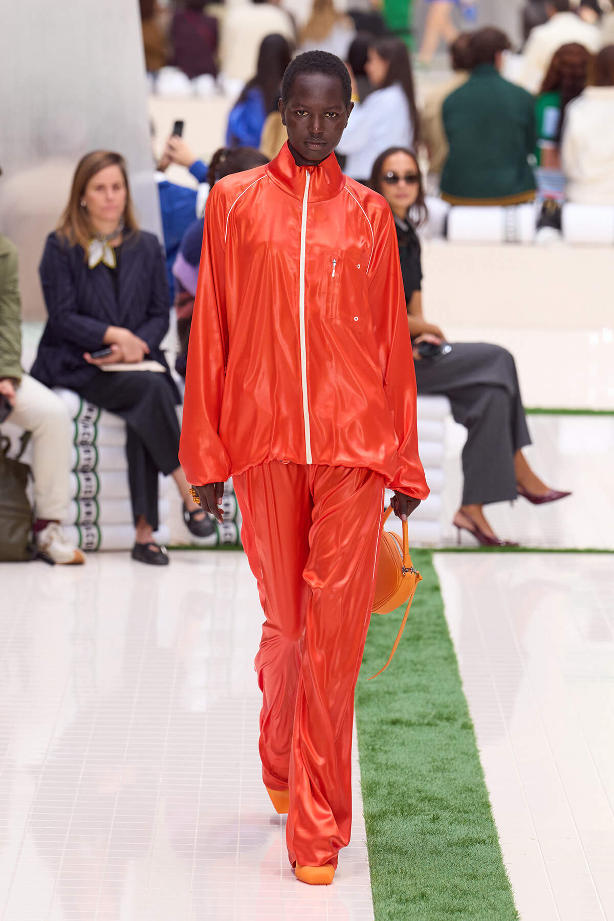 Model on a fashion runway wearing an orange shiny tracksuit and carrying a small orange purse/messenger bag.