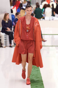 Male model walking a runway in a burnt orange hooded coat, matching shorts, and red sneakers during a fashion show with spectators in the background