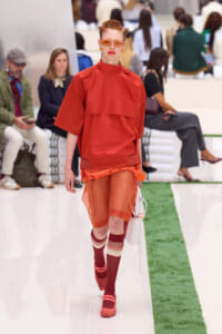 Model walking the runway in a bright orange monochrome outfit, red-framed sunglasses, and matching accessories with a white handbag.