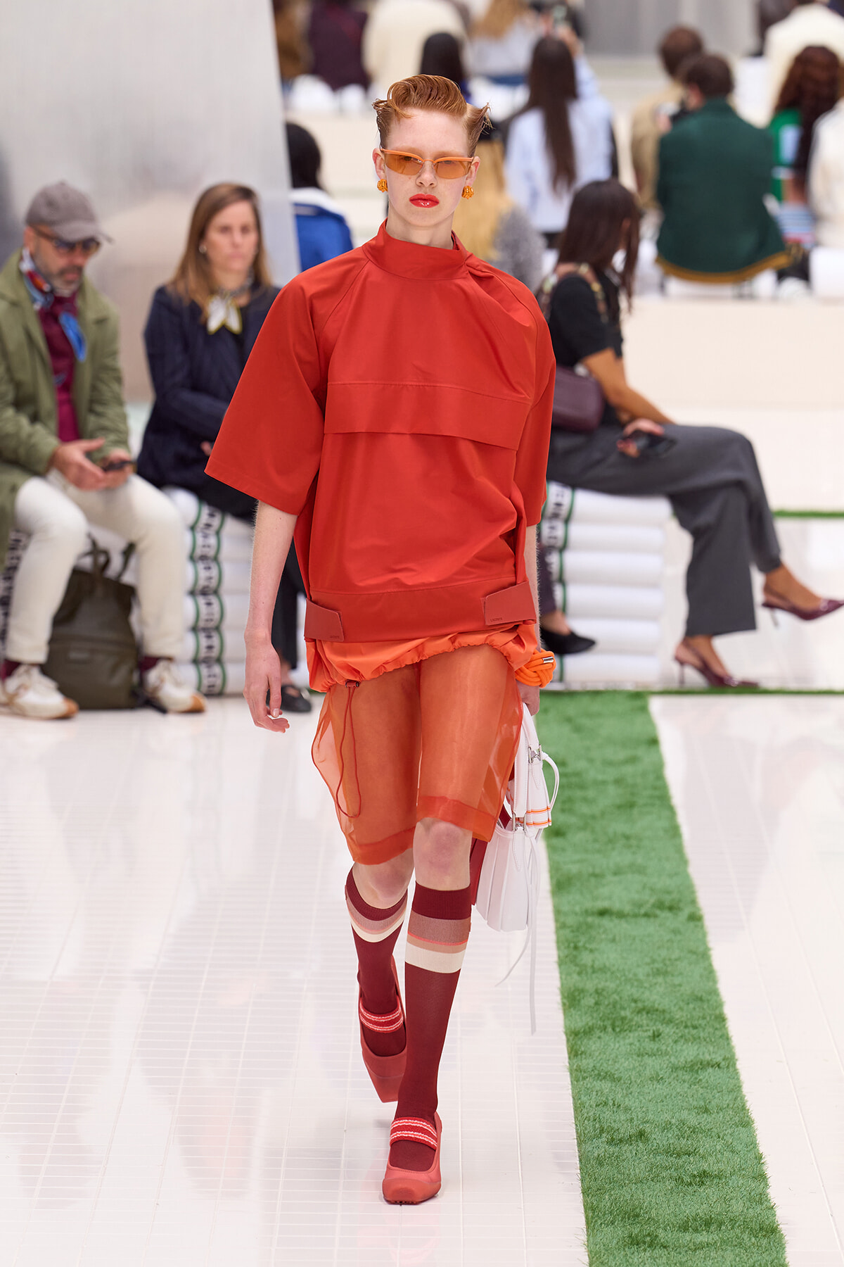 Model walking the runway in a bright orange monochrome outfit, red-framed sunglasses, and matching accessories with a white handbag.