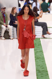 Model walking a runway in an orange sleeveless romper with matching sandals and orange accessories.