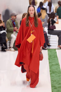 Model walks the runway in a bold red cape outfit with an orange pouch and layered necklaces, background spectators blurred.