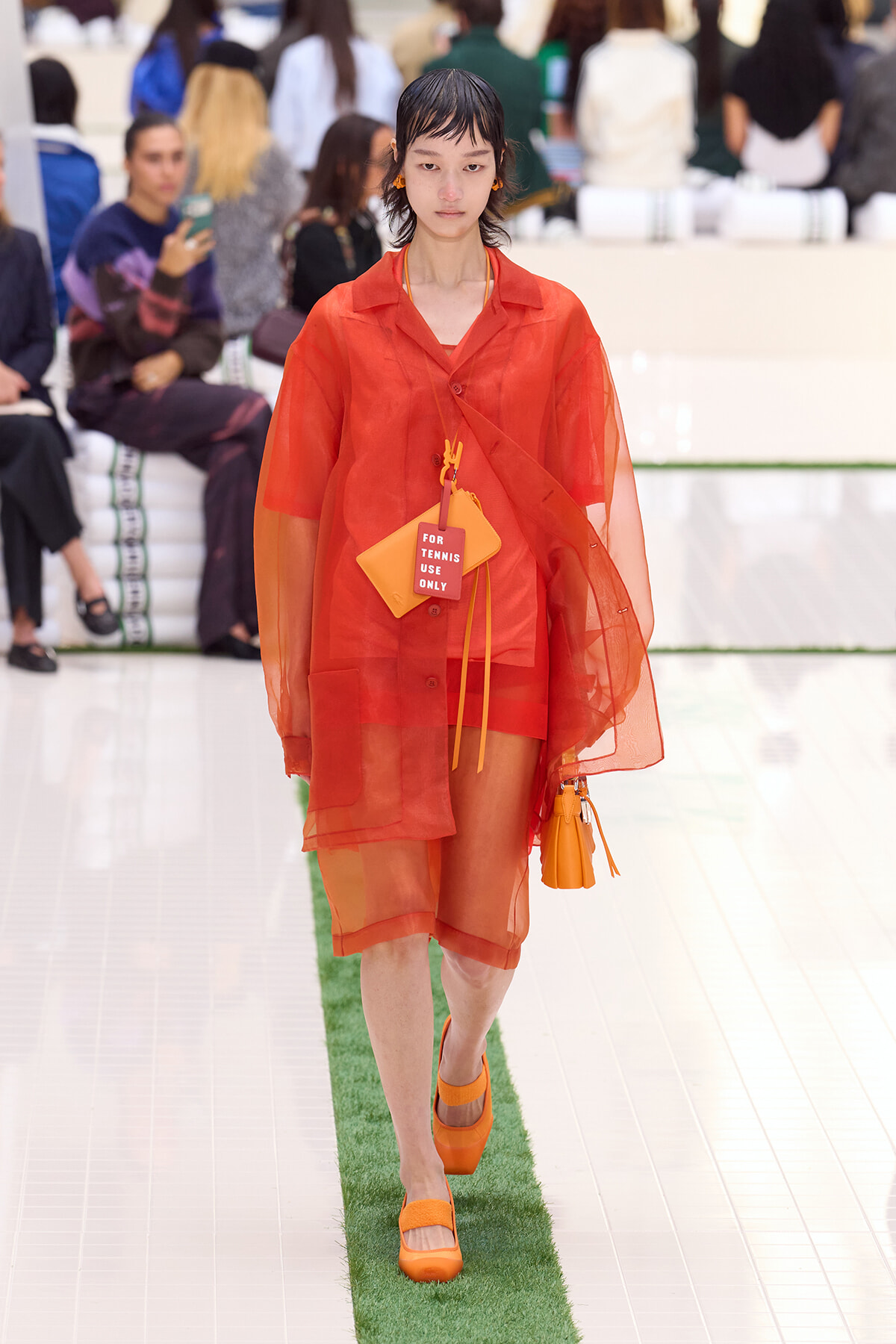Model walks the runway in a translucent orange shirt-dress set with matching shorts and orange shoes, carrying orange accessories and a tag reading 'FOR TENNIS USE ONLY'.