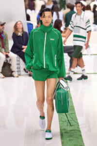 Male model in a bright green track suit walking a fashion show, carrying a matching green bag with white trim.
