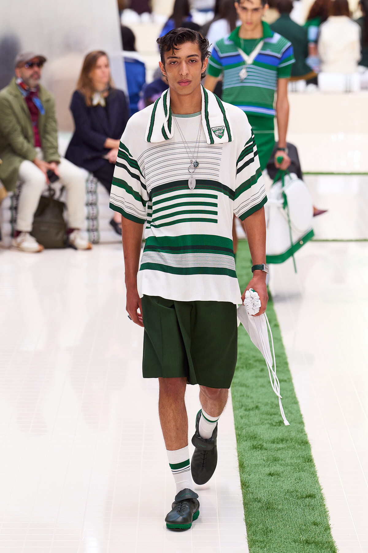 Male model walks a fashion show in a white and green striped polo, dark green shorts, white socks, and black shoes.