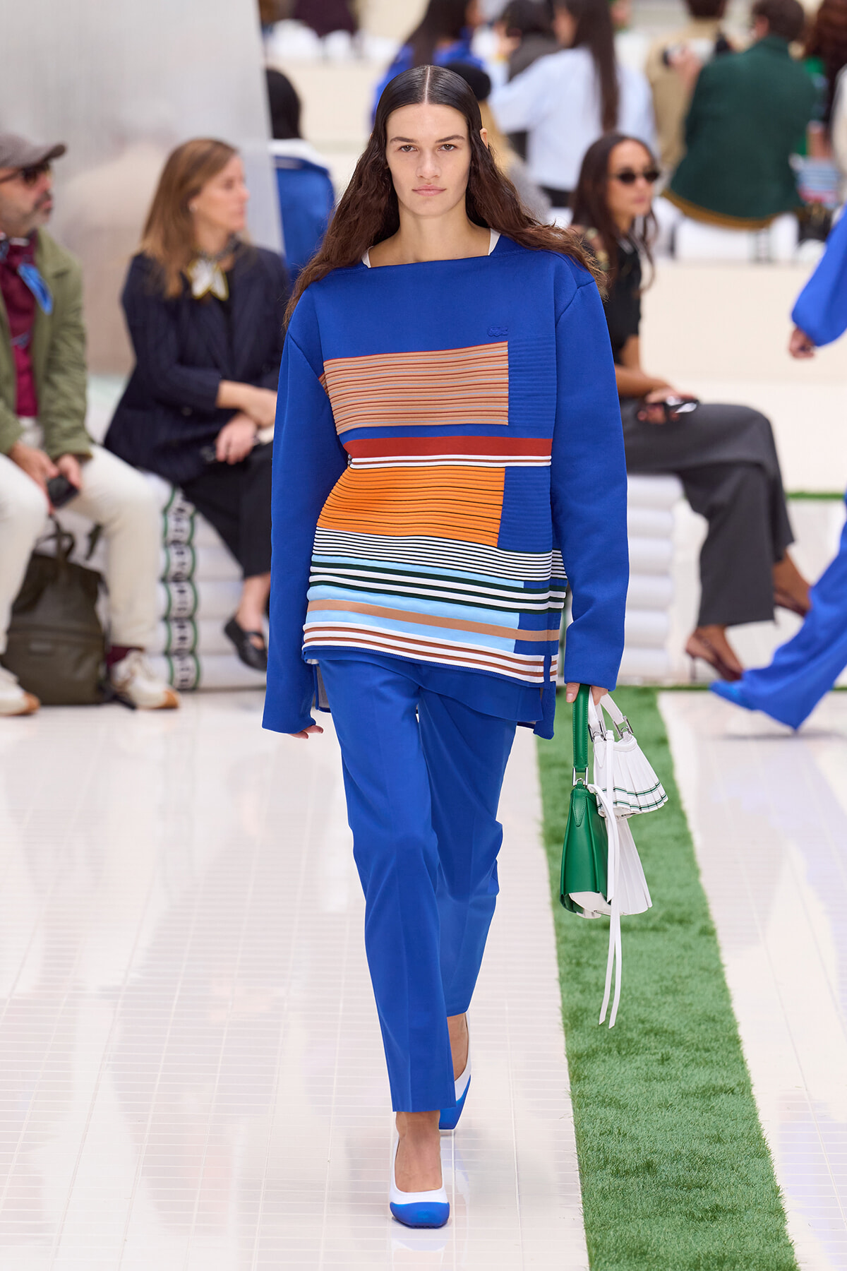 Model walking a runway in a cobalt blue ensemble with striped orange, white, and blue top; holds a green-and-white handbag.