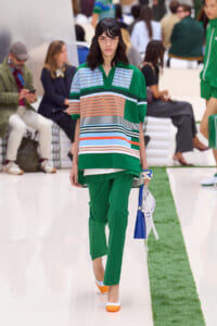 Runway model wearing a green striped polo over green pants, holding a white and blue handbag.