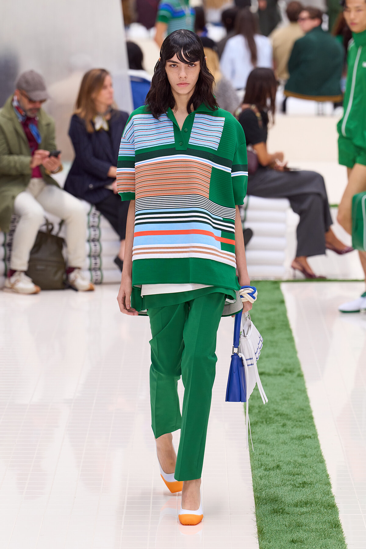 Runway model wearing a green striped polo over green pants, holding a white and blue handbag.