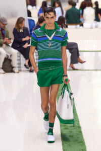 Male model walking a fashion show in a green striped polo and green shorts, carrying a white bag with green accents.