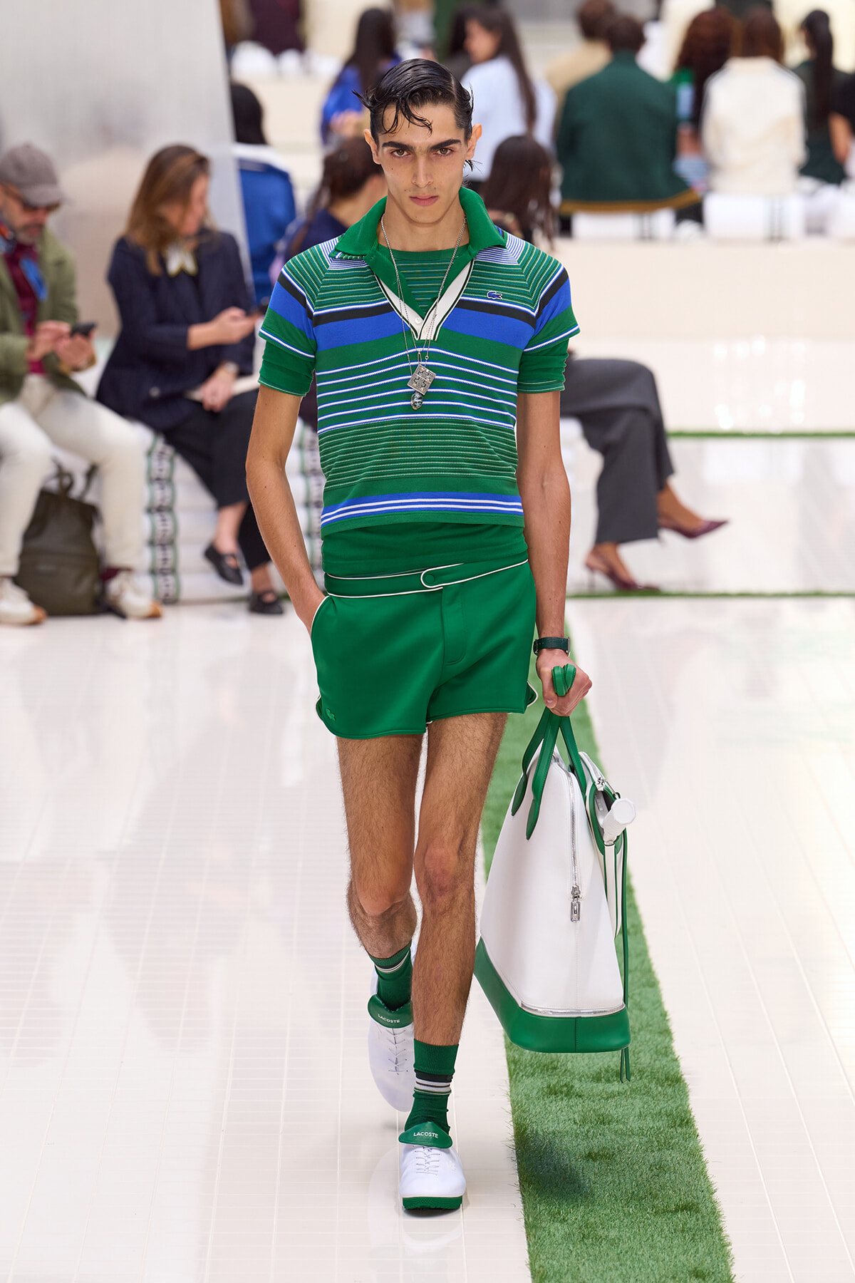Male model walking a fashion show in a green striped polo and green shorts, carrying a white bag with green accents.