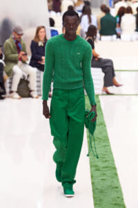Male model walking a runway in a monochrome green outfit: emerald knit sweater, matching green pants and shoes, holding a green accessory bag.