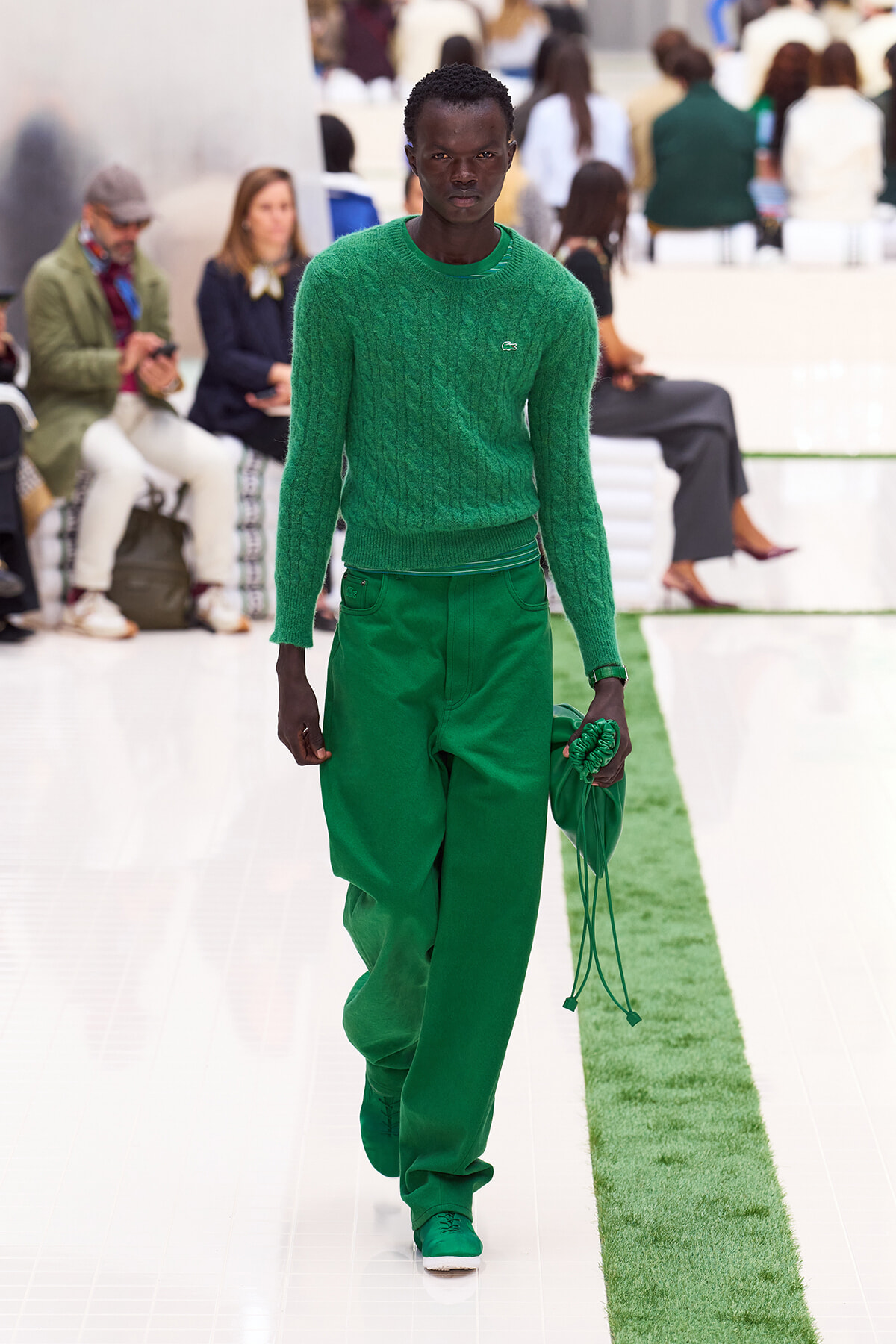 Male model walking a runway in a monochrome green outfit: emerald knit sweater, matching green pants and shoes, holding a green accessory bag.