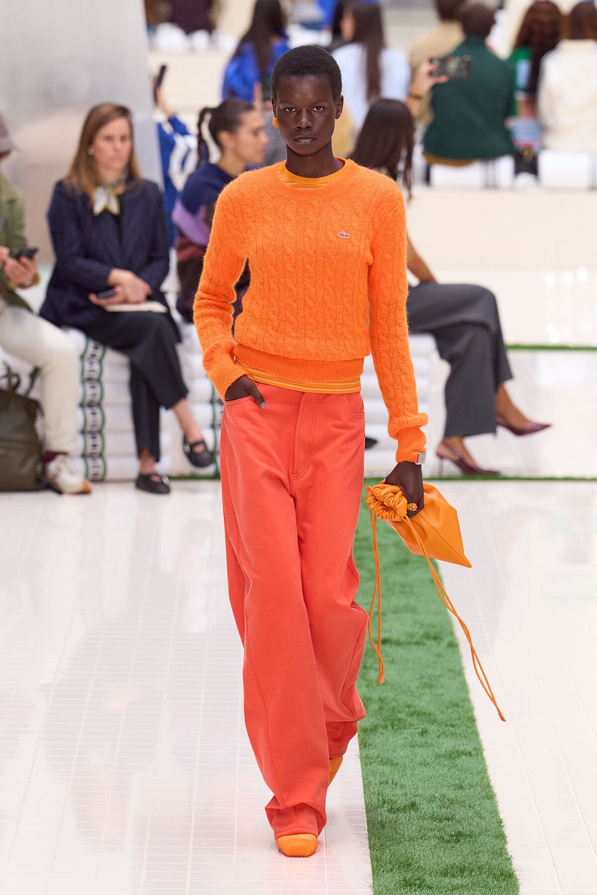 Model walks the runway in a bright orange cable-knit sweater and matching orange wide-leg pants, carrying an orange pouch bag.