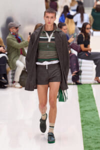 Male model walks the runway in a layered brown coat over a green striped top, gray shorts, and green shoes, carrying a green bag.