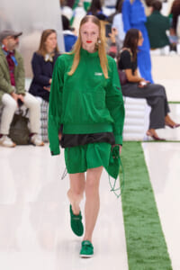 Model walks the runway in a green Lacoste tracksuit with matching sneakers and shorts at a fashion show, studio-style setting.